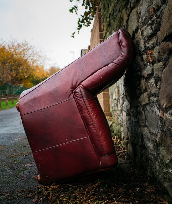 A large, red leather armchair has been positioned upside down and leaning against a stone wall on a residential pavement area. The armchair appears to be in the process of removal or transportation, w