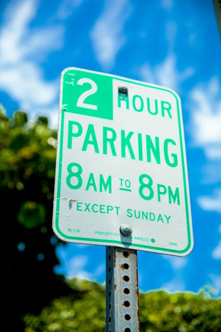 A close-up photograph of a green and white parking restriction sign mounted on a weathered metal pole, displaying a two-hour parking limit from 8 am to 8 pm, except Sundays, in Putney. The sign is positioned outdoors against a background of a bright blue sky with some white clouds and blurred green foliage, indicating a sunny day. In the context of house removals or relocations, this parking sign may be relevant for planning vehicle access and loading logistics on the day of a house move. The sign's clear textual instructions assist in understanding parking restrictions that could impact the scheduling of furniture transport or loading and unloading operations, which Putney Man and Van, a professional removals service, would consider during home relocation activities.