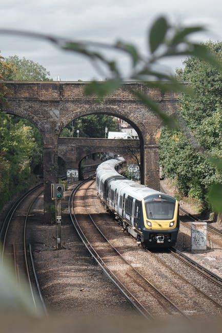 A modern passenger train with a sleek yellow and grey exterior is moving along railway tracks beneath an old brick railway bridge with three large archways, surrounded by lush green trees. The train is captured in motion, heading toward the viewer, with visible windows reflecting light. The railway tracks run parallel, with a signal light and a small electrical box nearby, indicating an active railway line. The scene is set outdoors during daytime, with overcast skies providing diffuse lighting. The image emphasizes railway infrastructure relevant to transportation and logistics associated with home relocation or moving services, such as those offered by Putney Man and Van, with visible elements of furniture and packing materials typically used in house removals, hinted at the context of moving objects via transport. The partial leaves in the foreground add depth to the composition and subtly frame the scene.