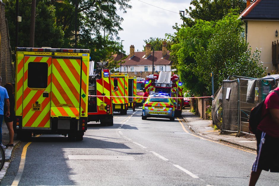 A street scene showing multiple emergency response vehicles, including a yellow London ambulance and several fire trucks with red and yellow chevron markings, parked along a residential area with trees and houses in the background. The vehicles are positioned across the road, partially blocking the street, with some personnel visible near the trucks. A police car with flashing lights is also present, and the area is cordoned off with red and white tape. On the right side, a woman in a pink top and black shorts is partially visible, standing behind a temporary fence. The scene suggests an active emergency or incident response in a residential neighbourhood, with lighting indicating daytime. The presence of emergency vehicles and personnel highlights the importance of safety and coordination during home relocation or emergency removal services, which Putney Man and Van can assist with for urgent situations.