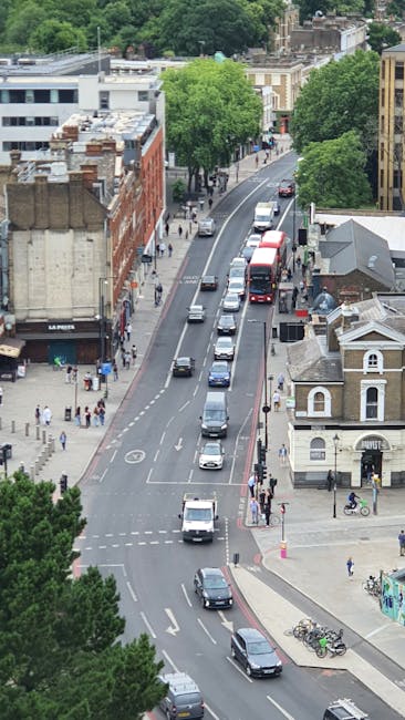 An aerial view of a city street during daytime showing a line of parked cars along the curb and moving vehicles including a black van and a red double-decker bus. Pedestrians are walking on the sidewalks, some near storefronts and buildings. To the right, a small white building with arched windows and a gabled roof is visible, alongside bicycles parked on the pavement. The street is lined with trees with lush green foliage, and behind them are mixed-use buildings, including brick and modern structures. The scene depicts an active urban environment suitable for house removals and furniture transport logistics, as part of a home relocation process. The photograph captures the loading process of household items in a busy city setting, consistent with the services provided by Putney Man and Van for removals.