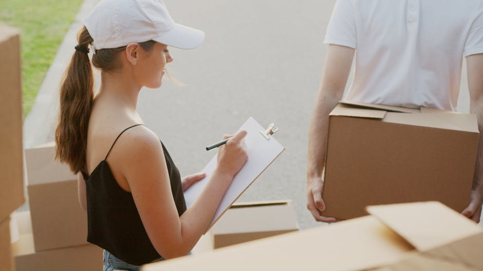 A young woman with long brown hair tied back and wearing a white cap, black sleeveless top, and light-colored pants, is holding a clipboard with a pen and appears to be taking notes. She is standing outdoors beside a stack of cardboard boxes on a pavement next to a doorway. A person with a white t-shirt is partially visible, holding a medium-sized closed cardboard box. The scene is well-lit with natural daylight, and the background includes a partly visible outdoor area, likely representing a home or property where furniture and household items are being prepared for home relocation or removal. This image depicts part of the packing and loading process linked to moving services, with objects and packing materials arranged around them. The professional removal process observed aligns with services offered by Putney Man and Van, highlighting furniture transport and loading activities.