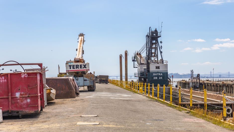 A view of a loading bay near Putney Pier during a home relocation process, showing a paved area with various equipment and vehicles used for furniture transport and packing. On the left side, there are large red and yellow containers, likely for storing packing materials or waste. In the center, a white crane labeled 'TEREX' is positioned on the paved surface, possibly used for lifting heavy furniture boxes or appliances. To the right, a large industrial crane labeled 'DUTRA DB 24' is situated near the water, with scaffolding and equipment for loading or unloading goods from boats or ships, indicating proximity to the river. In the background, there are additional vehicles and structures, with a view of the water extending towards the horizon under a partly cloudy sky. The scene captures the logistics involved in a professional removals or house moving service, emphasizing the loading, transport, and packing processes related to residential relocations near Putney Pier.