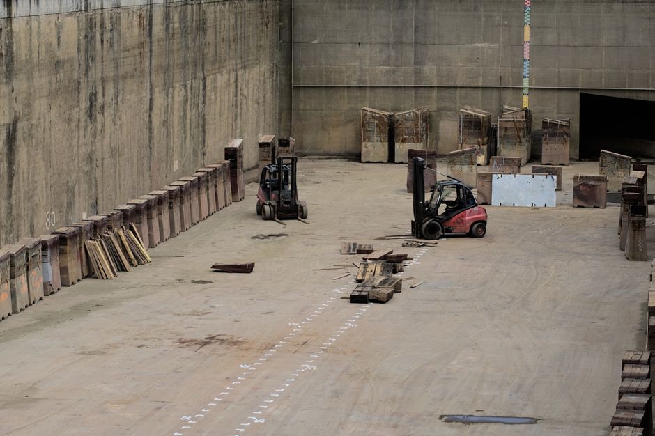 Inside a large, industrial loading bay with high concrete walls, two red and black forklifts are positioned on a cement floor, seemingly preparing for furniture or boxes to be moved. The floor has markings and scattered wooden pallets, some stacked or laid flat, used for transporting heavy items during home relocation or furniture transport processes. Along the left wall, a row of large, metal shipping or packing containers with attached, angled metal panels is visible, likely used for secure packaging of household belongings. Towards the back of the bay, several sturdy, rusted metal crates or containers are lined up, some open or partially obscured. A few wooden planks, small wooden blocks, and packing materials are arranged on the floor near the forklifts. The environment suggests a setting for loading or unloading furniture and packed items during a professional removal or moving service. Occasionally, Putney Man and Van's name can be subtly associated with the scene as part of operational logistics for house removals near Putney Pier, highlighting the importance of organized logistics, packing, and safe transport in home relocation tasks.