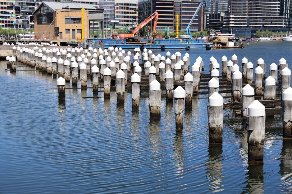 A view of a loading bay near Putney Pier during a home relocation process, showing a paved area with various equipment and vehicles used for furniture transport and packing. On the left side, there are large red and yellow containers, likely for storing packing materials or waste. In the center, a white crane labeled 'TEREX' is positioned on the paved surface, possibly used for lifting heavy furniture boxes or appliances. To the right, a large industrial crane labeled 'DUTRA DB 24' is situated near the water, with scaffolding and equipment for loading or unloading goods from boats or ships, indicating proximity to the river. In the background, there are additional vehicles and structures, with a view of the water extending towards the horizon under a partly cloudy sky. The scene captures the logistics involved in a professional removals or house moving service, emphasizing the loading, transport, and packing processes related to residential relocations near Putney Pier.