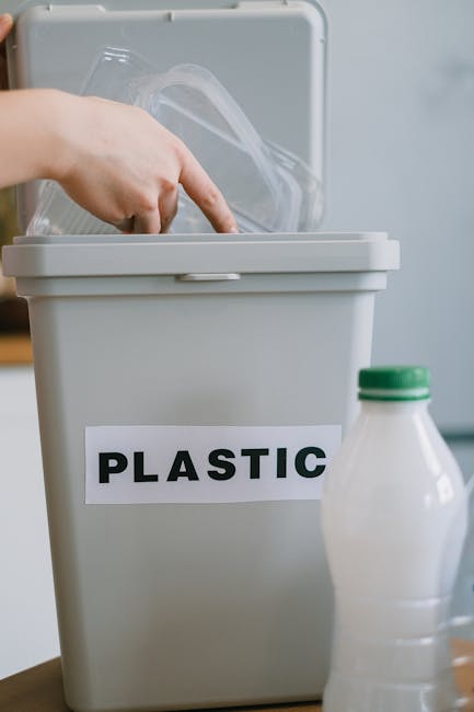 A white plastic recycling bin with a lid being opened by a person's hand, situated indoors. The bin has a label reading 'PLASTIC' in bold black letters on the front. Next to the bin, there is a plastic bottle with a green cap, possibly intended for recycling or disposal. The scene suggests an activity related to sorting waste materials, which is part of the home relocation process, involving packing and organizing recyclable items before moving. The background shows a neutral-colored wall, and the lighting is natural, providing clear visibility of the objects and their arrangement, consistent with a professional move or disposal service by Putney Man and Van.
