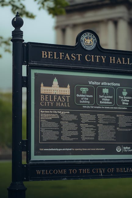 Close-up of a dark metal Belfast City Hall informational sign situated outdoors on a metal pole, with a background of green trees and a cloudy sky. The sign displays the Belfast City Hall logo, a detailed illustration of the building, and various visitor attraction icons, including guided tours, self-guided visitor exhibitions, and the Bobbin Coffee Shop. Below, it contains a lengthy section of text with bye-laws for City Hall grounds in white font on a dark background. The sign is part of the urban environment, often seen during home relocation or moving processes when residents or businesses are accessing city facilities for arrangements related to removals or disposal, reflecting the logistical considerations surrounding house moves or furniture transport within Belfast.