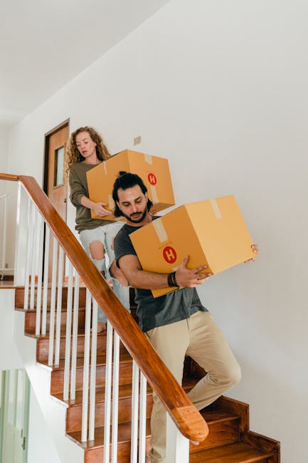 A man and a woman are carrying cardboard boxes during a home relocation inside a house with wooden staircase steps and a white painted interior wall. The man, dressed in a dark t-shirt and beige trousers, is positioned on the staircase climbing downward while holding a large cardboard box with packing tape and a red 'H' symbol, supporting it with both hands. The woman, wearing a brown jacket and ripped jeans, is walking behind him on the stairs, assisting with the boxes. The staircase has a wooden handrail and vertical balusters, and there is a door visible in the background. Natural light illuminates the scene, highlighting the furniture and packing materials indicating a typical moving process. This image exemplifies furniture transport and packing during a house move, as handled by Putney Man and Van, specializing in removals and staircases and narrow-access moves in Putney.