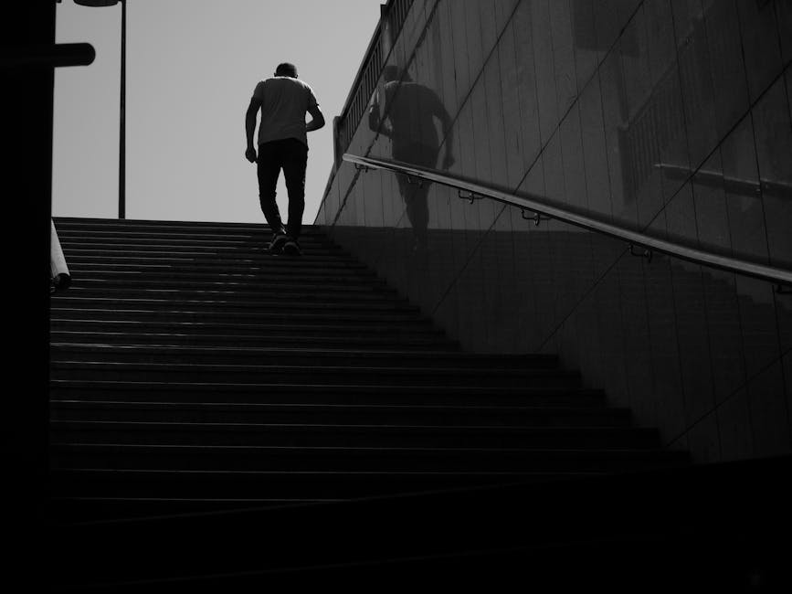 A person ascending an outdoor staircase made of flat, dark-colored steps with a metal handrail on the right side. The individual is dressed in casual clothing and is carrying a bag or object in their right hand. Their shadow is cast on the adjacent reflective wall, which mirrors the surroundings. The scene appears to be in an urban environment, possibly near a modern building with sleek exterior surfaces. The lighting is high contrast, emphasizing the silhouette of the person and the structure of the staircase, which may be part of a home relocation or moving process. The overall setting suggests a moment during furniture transport or packing and moving routines managed by Putney Man and Van, who specialise in removals, including navigating narrow or challenging access points.