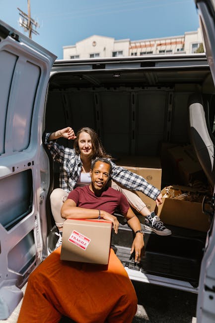 Two women engaged in a home relocation process are pictured at the rear of a large moving van, which is parked outdoors close to residential buildings. One woman, seated on the edge of the van's loading area, holds a medium-sized cardboard box marked 'FRAGILE' in red letters, while the other woman stands behind her, smiling and leaning inside the vehicle with her hand on her hip. The interior of the van contains additional cardboard boxes, some wrapped in protective plastic or packing paper, indicating careful packing. The women are dressed casually, with one wearing a red t-shirt and the other in a checkered shirt, suitable for a furniture transport and packing and moving service. The loading area is well-lit by natural daylight, and the scene reflects the process of loading household items for a house move, with cardboard boxes and packing materials prepared for transport. This image exemplifies actions typical during a professional removals service by Putney Man and Van, focusing on home relocation logistics and furniture transportation.
