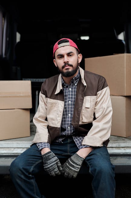 A man with a serious expression, wearing a light grey beanie, a green and navy puffer vest over a navy jacket, stands inside an open moving van. Behind him, cardboard moving boxes are stacked on the vehicle's flooring, ready for a house relocation. The van's interior is visible, with dark curtains and practical equipment for furniture transport. Outside the vehicle, the surrounding environment suggests a residential area. This scene depicts the loading process involved in packing and moving services provided by Putney Man and Van, highlighting preparations for same-day house removals in Putney, SW15, with professional logistical support for efficient furniture transport and packing throughout the home relocation process.