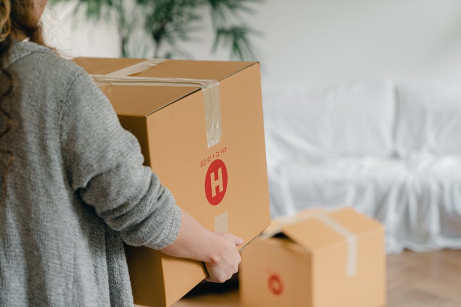 A person wearing a grey sweater is preparing to move large cardboard boxes in an indoor setting as part of a home relocation process. The individual is holding a knife, sealing the top of a medium-sized box marked with a red 'H' logo and printed details, indicating it contains household items. Behind them, there are other similarly packed cardboard boxes, some placed on the wooden floor, reflecting an organized packing and moving activity. The background features a white sofa with a fabric slipcover and some greenery, suggesting the interior of a residential property. The scene is well-lit with natural light, emphasizing the careful handling and packing procedures involved in furniture transport and house removals, with the boxes ready for loading into a van or vehicle by Putney Man and Van, specialists in removals and relocation services available at https://putneymanandvan.org.uk.