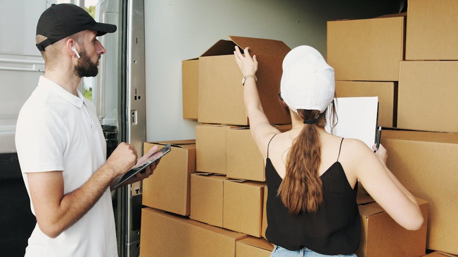 A person wearing a grey sweater is preparing to move large cardboard boxes in an indoor setting as part of a home relocation process. The individual is holding a knife, sealing the top of a medium-sized box marked with a red 'H' logo and printed details, indicating it contains household items. Behind them, there are other similarly packed cardboard boxes, some placed on the wooden floor, reflecting an organized packing and moving activity. The background features a white sofa with a fabric slipcover and some greenery, suggesting the interior of a residential property. The scene is well-lit with natural light, emphasizing the careful handling and packing procedures involved in furniture transport and house removals, with the boxes ready for loading into a van or vehicle by Putney Man and Van, specialists in removals and relocation services available at https://putneymanandvan.org.uk.