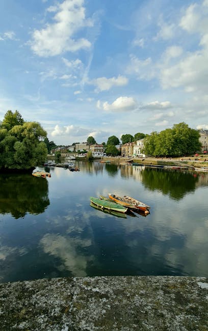 A scenic view of a calm river reflecting a blue sky with scattered white clouds. Along the riverbank, there are several trees with lush green foliage, and a row of residential buildings with light-colored facades and flat roofs. In the foreground, there are a few small wooden boats and one green boat moored close to the water's edge. The scene is captured during daylight, with natural lighting illuminating the river and surrounding landscape. This image depicts a peaceful riverside environment, with potential relevance to home relocation or moving services like those offered by Putney Man and Van, as they facilitate furniture transport and packing during house removals.