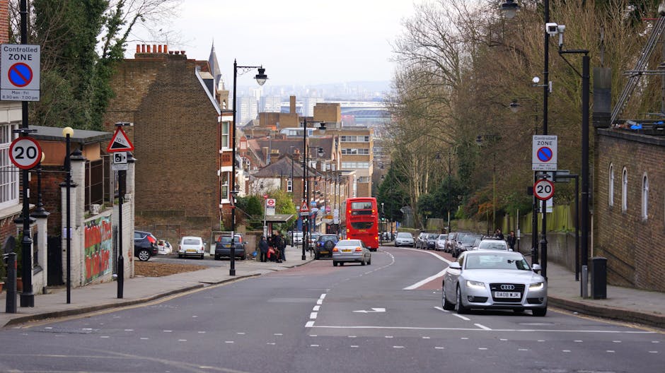 A street scene on Upper Richmond Road showing a residential area with brick houses and trees on both sides, alongside parked cars lining the curb. In the foreground, a silver Audi is parked on the right side, while a red double-decker bus is arriving or departing further down the road. Lamp posts and street signage, including a no-parking zone and speed limit signs, are visible, with some signage indicating controlled parking zones. The street appears to be part of a home relocation or moving process, with the environment well-lit by daytime natural light. In the background, additional vehicles and urban buildings can be seen, highlighting a typical busy neighbourhood in Putney. This scene illustrates a typical setting where Putney Man and Van might be engaged in furniture transport or house removals, with an emphasis on the street's layout, parked vehicles, and the general movement of traffic and pedestrians related to moving services.